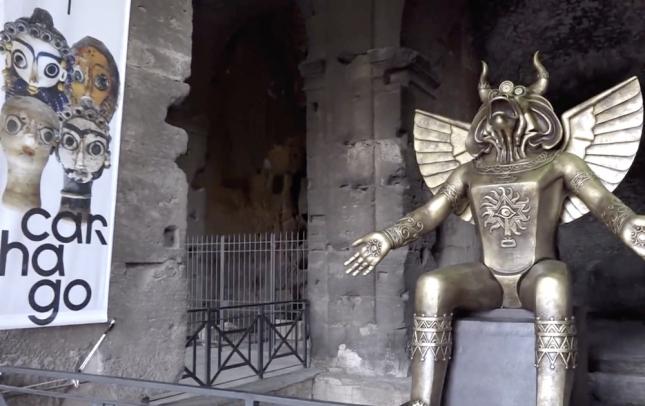 O deus pagão Moloque posta ao lado de uma placa de exposição no Coliseu, Roma, 27 de setembro de 2019.sanmarinotv.sm/ screen grab